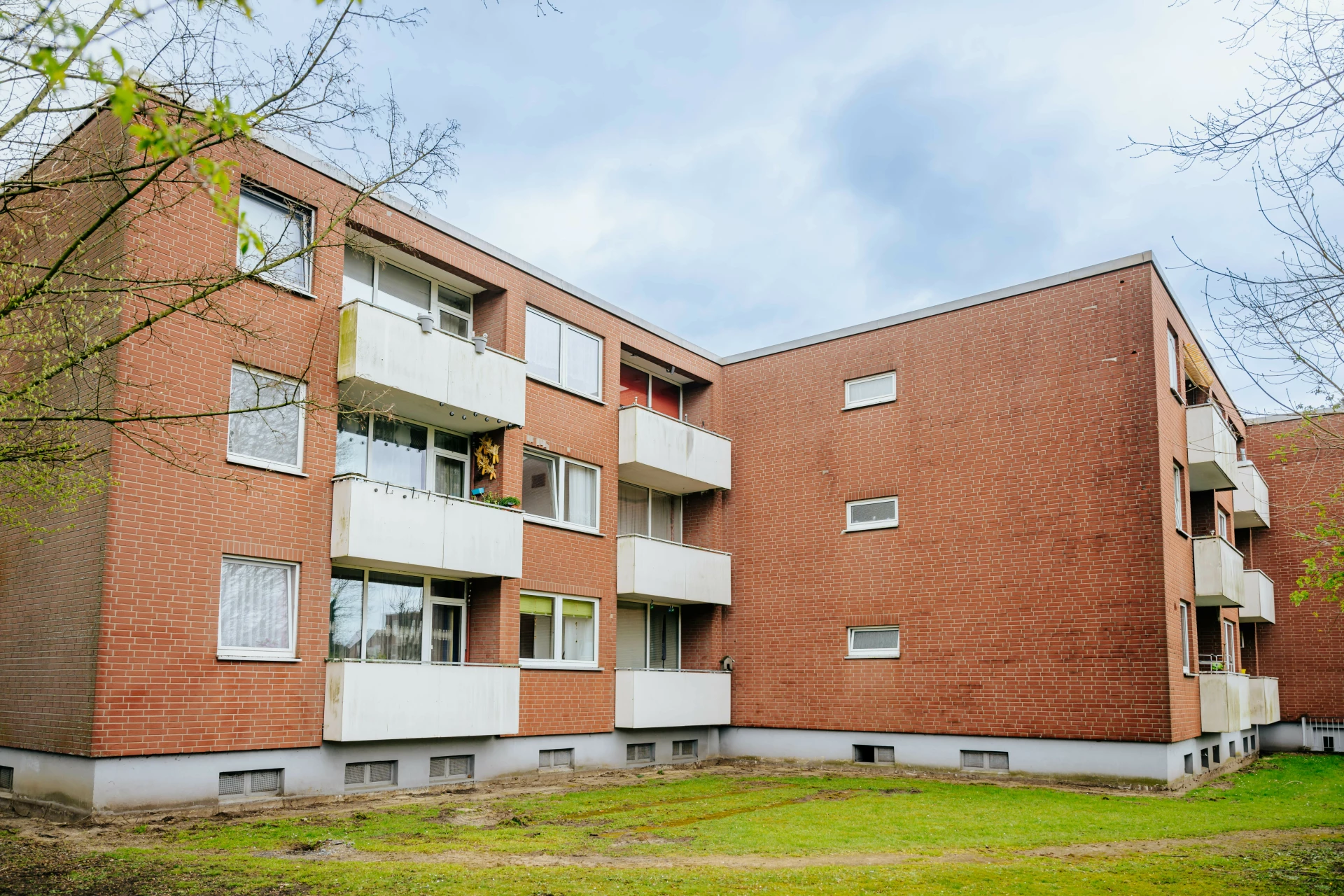 Bestandsaufnahme eines Mehrfamilienhauses vor der Modernisierung. Das Gebäude hat eine rote Klinkerfassade, mehrere Etagen und teilweise vorhandene Balkone. Es ist von einer Rasenfläche und einzelnen Bäumen umgeben und zeigt den baulichen Zustand vor geplanten Modernisierungsmaßnahmen.