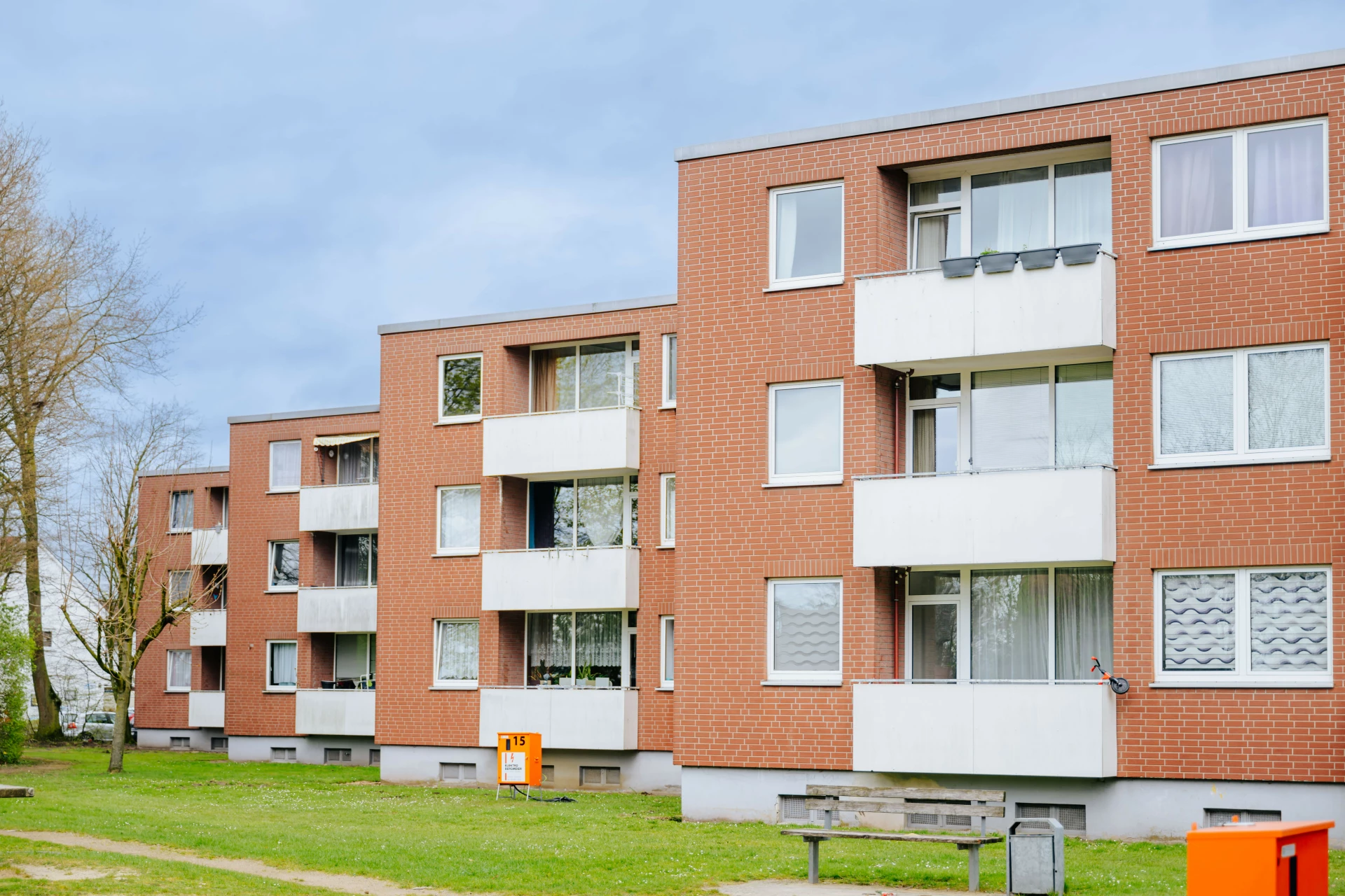 Mehrgeschossiges Wohngebäude aus rotem Backstein mit weißen Balkonbrüstungen. Mehrere Wohnungen verfügen über angesetzte Balkone, die gleichmäßig an der Fassade angeordnet sind. Vor dem Haus erstreckt sich eine gepflegte Grünfläche mit Rasen, Sitzbank und Abfallbehältern. Die Gebäude stehen in einer ruhigen Wohnlage mit Bäumen und viel Tageslicht. Das Erscheinungsbild wirkt ordentlich und funktional.
