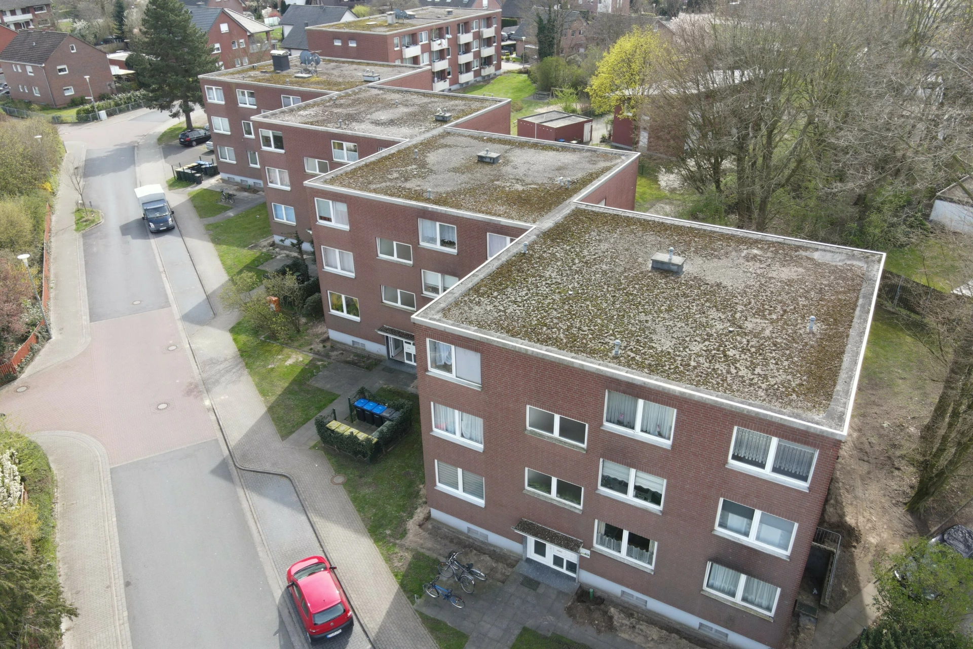 Luftaufnahme mehrerer Mehrfamilienhäuser mit roter Klinkerfassade und Flachdächern. Die Gebäude stehen entlang einer Wohnstraße und sind von Grünflächen, Bäumen und Gehwegen umgeben. Parkende Autos sind vor den Häusern zu sehen. Die Aufnahme zeigt den baulichen Bestand eines Wohnquartiers aus der Vogelperspektive.