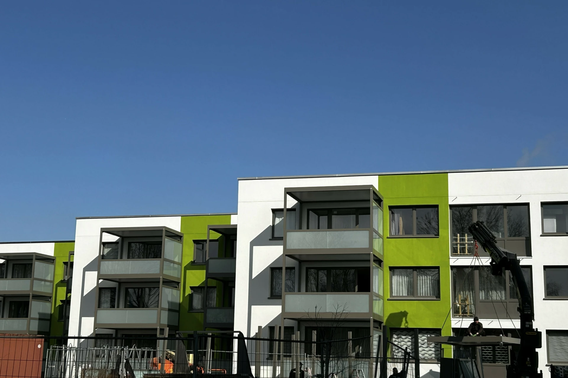 Auf dem Bild sind moderne Mehrfamilienhäuser mit weißen und grünen Fassaden zu sehen. Mehreren Balkone und klare Architektur vor blauem Himmel. Ein Baukran weist auf ein Neubau‑Wohnprojekt.