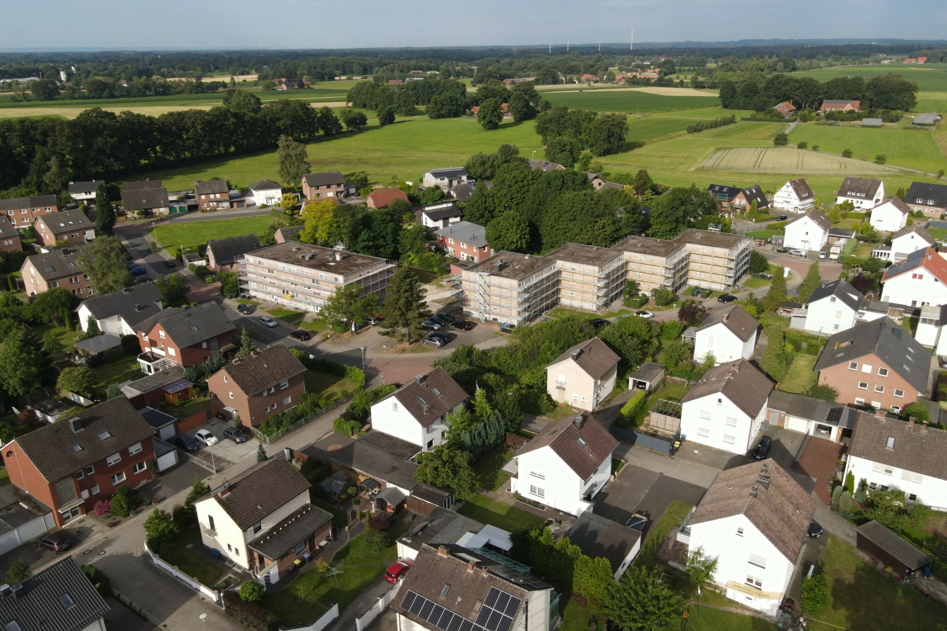 Luftaufnahme eines gewachsenen Wohnquartiers mit mehreren mehrgeschossigen Wohngebäuden und Einfamilienhäusern. Die Mehrfamilienhäuser liegen eingebettet zwischen grünen Gärten, Bäumen und Wohnstraßen. Die Gebäude haben helle Fassaden und sind gleichmäßig im Quartier verteilt. Im Hintergrund erstrecken sich offene Felder und landwirtschaftliche Flächen, die den ländlich‑ruhigen Charakter der Umgebung unterstreichen. Das Bild zeigt den aktuellen Bestand und das Umfeld der Wohnanlage vor einer geplanten Modernisierung.