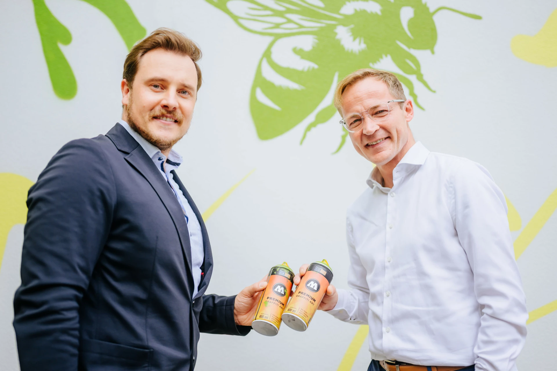 Zwei Männer stehen nebeneinander vor einer hellen Wand mit grüner, grafischer Naturillustration. Beide lächeln in die Kamera und halten jeweils zwei Getränkedosen in der Hand. Links trägt ein Mann ein dunkles Sakko mit hellem Hemd, rechts steht ein Mann in weißem Hemd. Die Szene wirkt freundlich, professionell und partnerschaftlich.