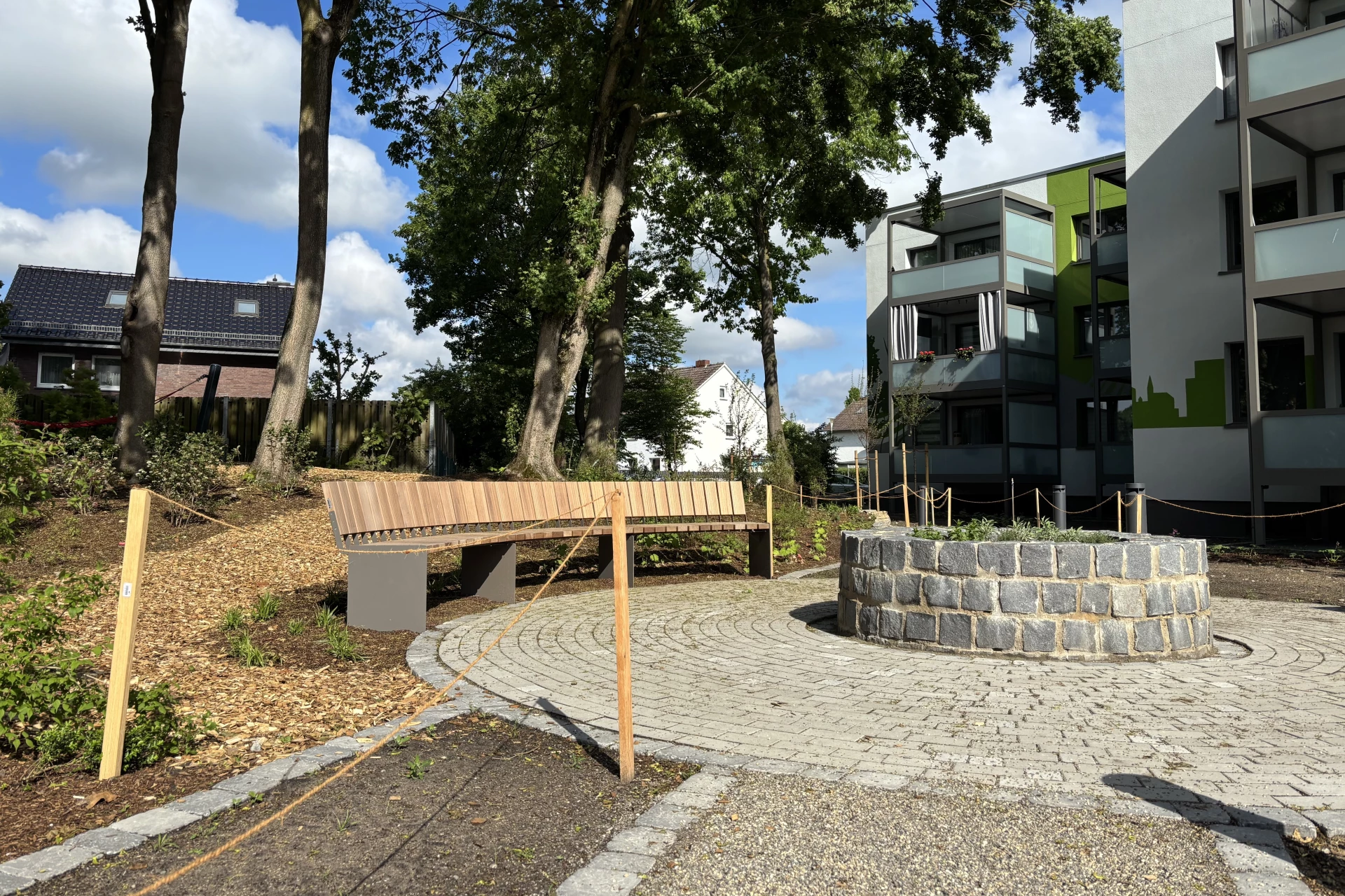 Das Bild zeigt einen neu gestalteten Außenbereich eines Wohnquartiers. Im Mittelpunkt befindet sich ein runder, gepflasterter Platz mit einer gemauerten Feuerstelle. Drei gebogene Holzbänke sind kreisförmig um den Platz angeordnet und laden zum Verweilen ein. Mehrere befestigte Wege führen auf den Sitzbereich zu. Im Hintergrund sind mehrgeschossige Wohngebäude zu sehen, darunter ein Haus mit einem großflächigen grünen Wandgemälde. Bäume mit frischem Laub und offene Grünflächen umrahmen den Platz. Der Bereich wirkt modern, offen und als gemeinschaftlicher Treffpunkt für Bewohnerinnen und Bewohner gestaltet.